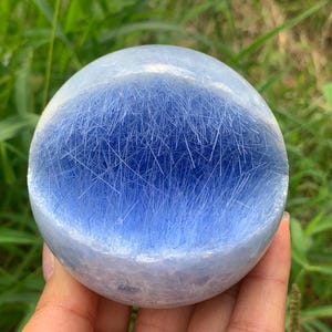 May include: A polished spherical gemstone featuring a translucent light blue body with a dense internal network of fine, needle-like white inclusions that create a fibrous, radiating pattern throughout the centre of the smooth, rounded stone.