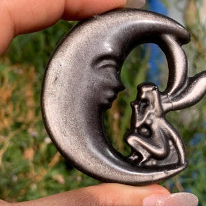May include: A crescent moon carving made of metallic silver-toned obsidian stone. The moon features a carved face profile on the upper curve, with a small winged fairy figure sitting on the lower curve, both rendered in a smooth, reflective finish.