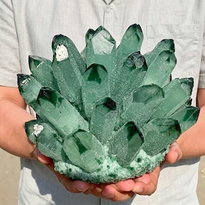 Pode incluir: Um grande aglomerado de cristais de quartzo verde-escuro com múltiplas pontas translúcidas que se elevam a partir de uma base sólida e texturizada. A formação mineral exibe vários tons de verde esmeralda e floresta com algumas inclusões brancas.