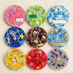 May include: Nine circular resin coasters featuring embedded colorful beads, charms, and custom name labels. Designs include floral, green, blue with rainbow, nautical, coffee, yellow with moon, red with flowers, and purple with emoji themes.