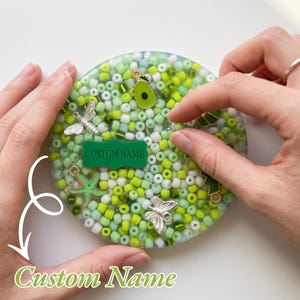 May include: A round, clear resin coaster filled with green and white beads. Silver charms include a dragonfly, starfish, and avocado. A green label with the text "CUSTOM NAME" is also present. The coaster is held by two hands.