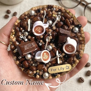 May include: A round, coffee-themed resin coaster with coffee beans, chocolate pieces, and miniature white coffee cups. Silver-colored cutlery and metallic beads are scattered throughout. The coaster has the text "Custom Name" and "For Lily".