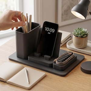 May include: A dark gray desk organizer featuring a vertical pen holder, a central smartphone stand displaying 10:09 AM, and a small tray for a coiled charging cable. The organizer is constructed from a matte material and sits on a flat base.