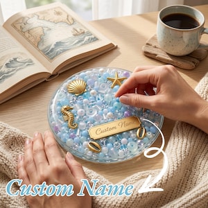 May include: A round, clear container filled with a mix of blue, white, and translucent beads, accented with gold-toned charms shaped like a starfish, seahorse, and seashells. A rectangular gold plaque in the center features the text Custom Name.