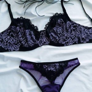 May include: Black and purple lingerie set featuring a bra and matching underwear. The bra has a floral lace design with scalloped edges. The underwear is a matching color and style. A black feather decoration is in the background.