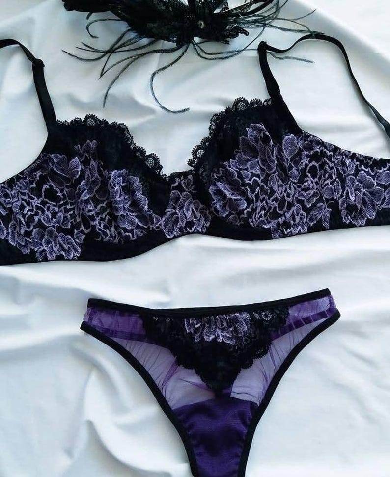 May include: Black and purple lingerie set featuring a bra and matching underwear. The bra has a floral lace design with scalloped edges. The underwear is a matching color and style. A black feather decoration is in the background.