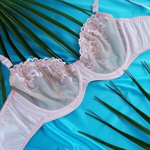 May include: A light pink bra with floral lace detailing on the cups. The bra has adjustable straps and a delicate, feminine design. The background is a vibrant blue fabric with green palm fronds.