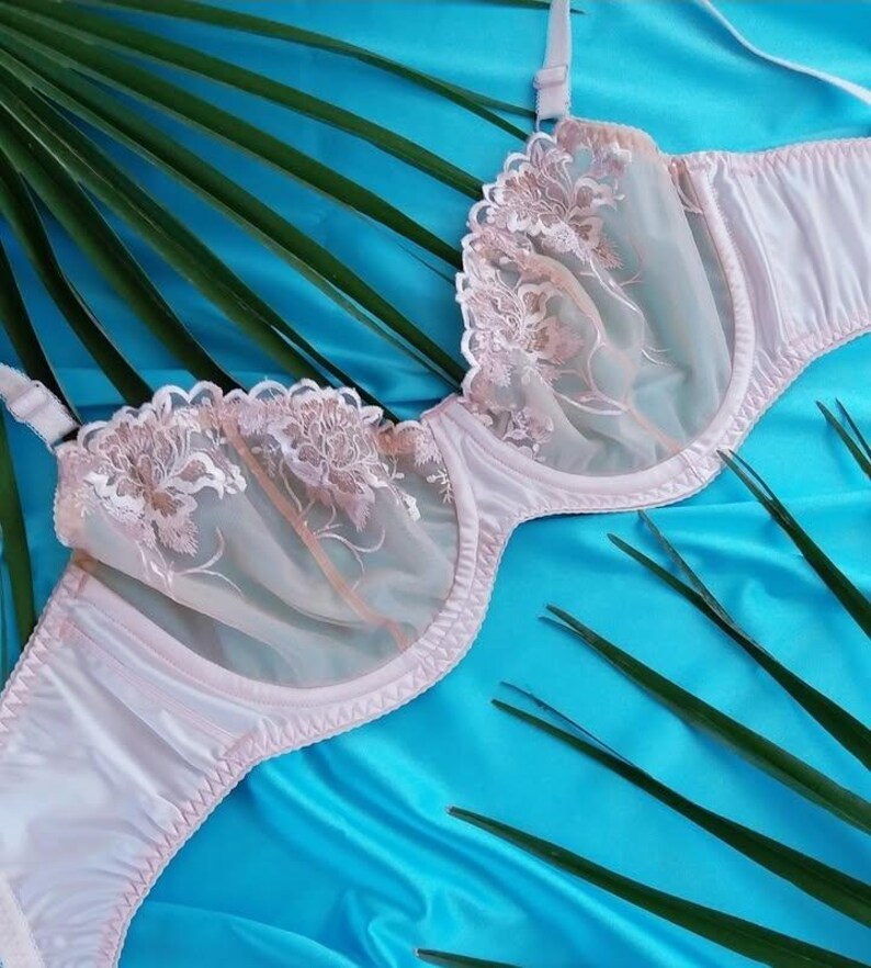 May include: A light pink bra with floral lace detailing on the cups. The bra has adjustable straps and a delicate, feminine design. The background is a vibrant blue fabric with green palm fronds.