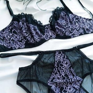 May include: Black lingerie set featuring a bra and matching underwear. The bra and underwear are adorned with a delicate floral lace pattern in shades of lavender and black. The set is made of sheer fabric.