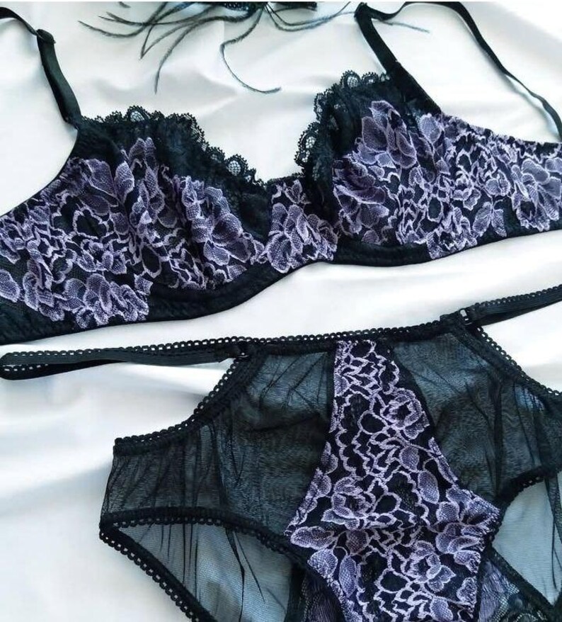May include: Black lingerie set featuring a bra and matching underwear. The bra and underwear are adorned with a delicate floral lace pattern in shades of lavender and black. The set is made of sheer fabric.