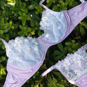 May include: A lavender bra and matching underwear set with delicate white floral lace detailing. The bra features a full coverage design with adjustable straps. The set is displayed on a bed of green leaves, creating a soft and feminine aesthetic.