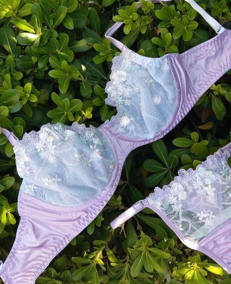 May include: A lavender bra and matching underwear set with delicate white floral lace detailing. The bra features a full coverage design with adjustable straps. The set is displayed on a bed of green leaves, creating a soft and feminine aesthetic.