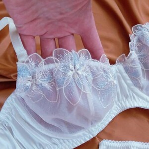 May include: White bra with floral embroidery in light blue and silver glitter. The bra features a satin finish and delicate lace detailing. The straps are white and adjustable. The image shows a close-up of the bra.