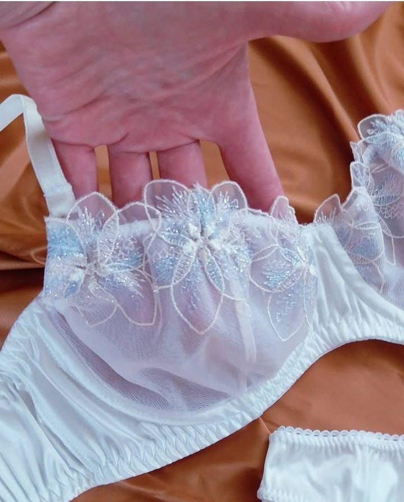 May include: White bra with floral embroidery in light blue and silver glitter. The bra features a satin finish and delicate lace detailing. The straps are white and adjustable. The image shows a close-up of the bra.