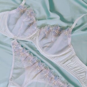 May include: White lingerie set featuring a bra and matching panties. The bra and panties are adorned with delicate floral lace in shades of blue and pink. The set is made of a soft, sheer fabric, creating a romantic and feminine aesthetic.