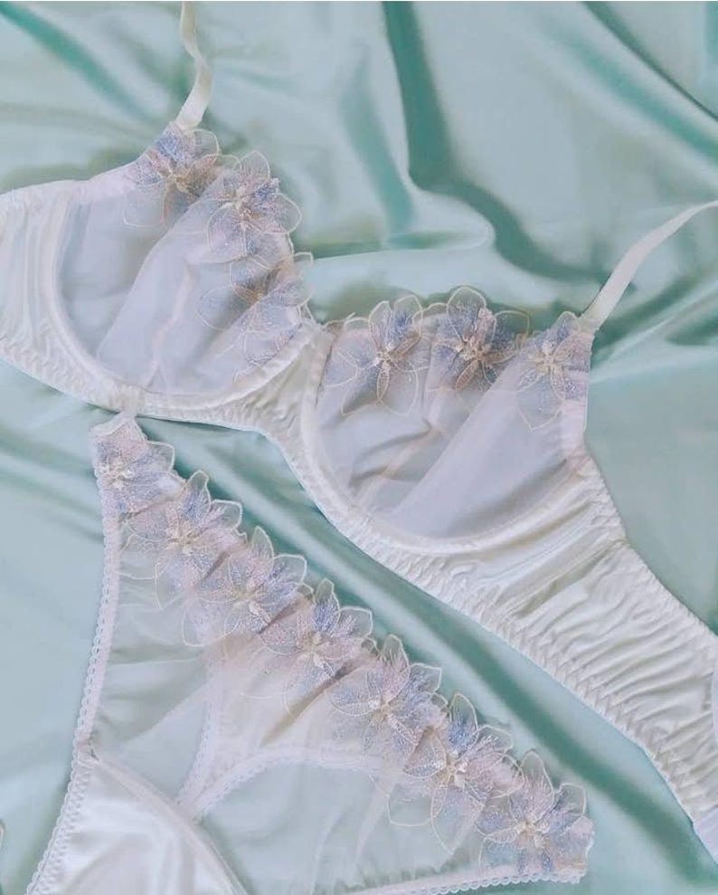 May include: White lingerie set featuring a bra and matching panties. The bra and panties are adorned with delicate floral lace in shades of blue and pink. The set is made of a soft, sheer fabric, creating a romantic and feminine aesthetic.