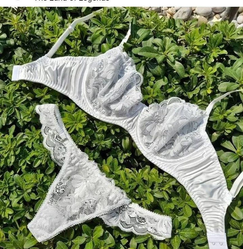 May include: A white lingerie set featuring a bra and matching thong. The bra has underwire support and lace detailing on the cups. The thong also has lace accents. The set is made of a smooth, satin-like material and is displayed on a bed of green foliage.
