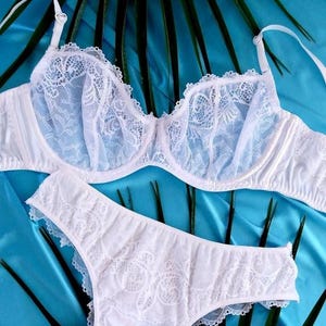 May include: White lingerie set featuring a bra and matching underwear. The bra has delicate lace detailing on the cups and straps. The underwear also has lace accents. The set is displayed on a blue background with green leaves.
