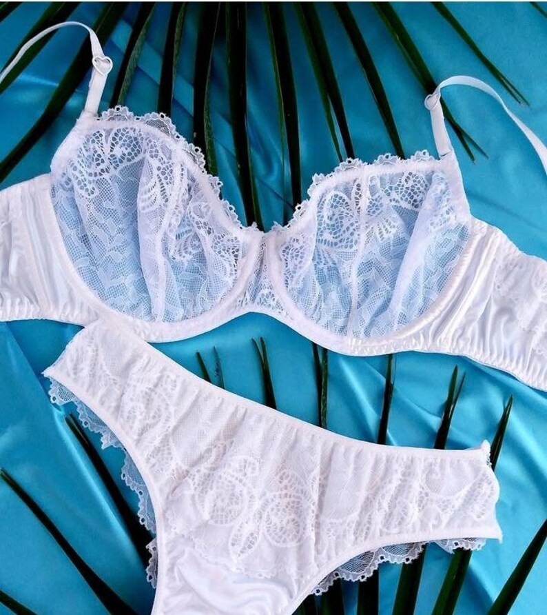 May include: White lingerie set featuring a bra and matching underwear. The bra has delicate lace detailing on the cups and straps. The underwear also has lace accents. The set is displayed on a blue background with green leaves.