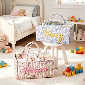 May include: Two fabric baby caddies with handles, one with a blue floral pattern and the name "Henry" in yellow, the other with a floral pattern and the name "Harper" in pink. Both are filled with baby supplies. Toys and a bed are in the background.