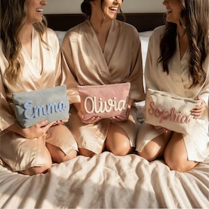 May include: Three corduroy cosmetic bags in pastel shades: blue, pink, and beige. Each bag features a name embroidered in a contrasting colour: "Emma", "Olivia", and "Sophia". The bags are held by people wearing matching silk robes.
