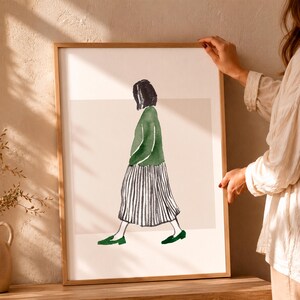 Könnte beinhalten: Ein gerahmter Kunstdruck mit einer Aquarellillustration einer Person mit kurzem dunklem Haar, die im Profil geht. Die Figur trägt ein grünes Langarmshirt, einen schwarz-weiß vertikal gestreiften Rock und grüne flache Schuhe.