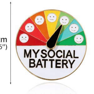 May include: A round enamel pin, 1.6 inches in diameter, with a gauge design labeled "MY SOCIAL BATTERY." The gauge features a needle and a color-coded scale from red (sad) to green (happy), with corresponding smiley faces.
