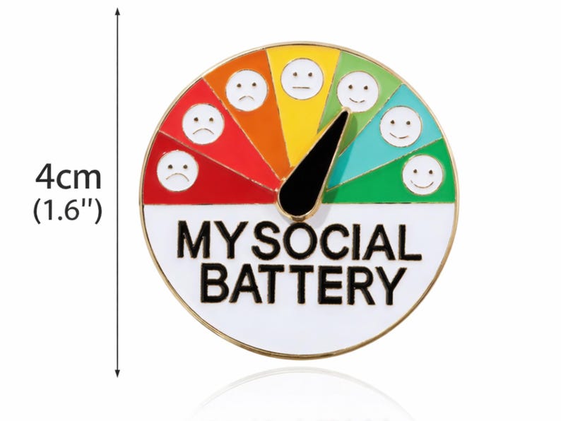 May include: A round enamel pin, 1.6 inches in diameter, with a gauge design labeled "MY SOCIAL BATTERY." The gauge features a needle and a color-coded scale from red (sad) to green (happy), with corresponding smiley faces.