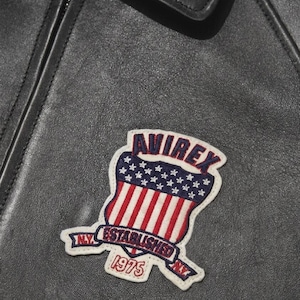 Puede incluir: Primer plano de una chaqueta de cuero negra con un parche. El parche presenta la palabra "AVIREX" sobre un dise&ntilde;o de escudo con rayas rojas y blancas y estrellas azules. Tambi&eacute;n se ve el texto "ESTABLISHED 1975".