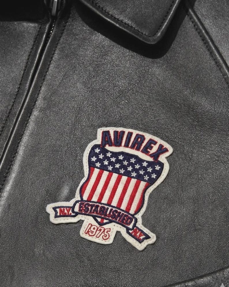 Puede incluir: Primer plano de una chaqueta de cuero negra con un parche. El parche presenta la palabra "AVIREX" sobre un dise&ntilde;o de escudo con rayas rojas y blancas y estrellas azules. Tambi&eacute;n se ve el texto "ESTABLISHED 1975".