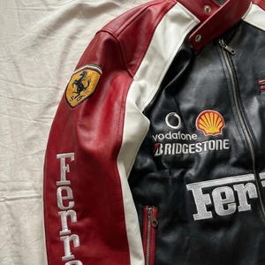 Op de afbeelding: Een zwarte, rode en witte leren jas met geborduurde Ferrari-branding. De jas heeft een Ferrari-logopatch op de linkermouw, sponsorlogo's en het woord "Ferrari" geborduurd op de linkermouw. De jas heeft een ritssluiting.