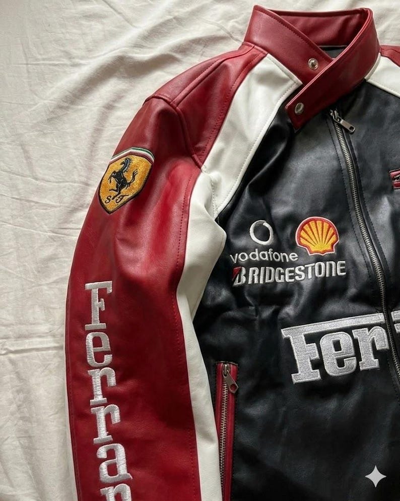 Op de afbeelding: Een zwarte, rode en witte leren jas met geborduurde Ferrari-branding. De jas heeft een Ferrari-logopatch op de linkermouw, sponsorlogo's en het woord "Ferrari" geborduurd op de linkermouw. De jas heeft een ritssluiting.