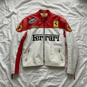 May include: A white and red Ferrari racing jacket with black lettering and logos. The jacket features a zipper closure, two front pockets, and patches including the Ferrari logo, Arai, and Michelin.