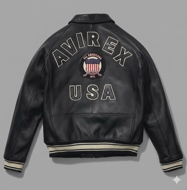 Puede incluir: Chaqueta de cuero negra con la palabra "AVIREX" en letras blancas en la espalda. Debajo, un parche con la bandera americana y la palabra "USA". La chaqueta tiene cuello y pu&ntilde;os de canal&eacute;.