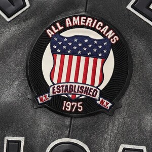 Puede incluir: Primer plano de una chaqueta de cuero negra con un parche circular. El parche presenta un dise&ntilde;o de escudo con rayas rojas y blancas, estrellas azules y las palabras "ALL AMERICANS ESTABLISHED 1975 N.Y." en letras blancas.