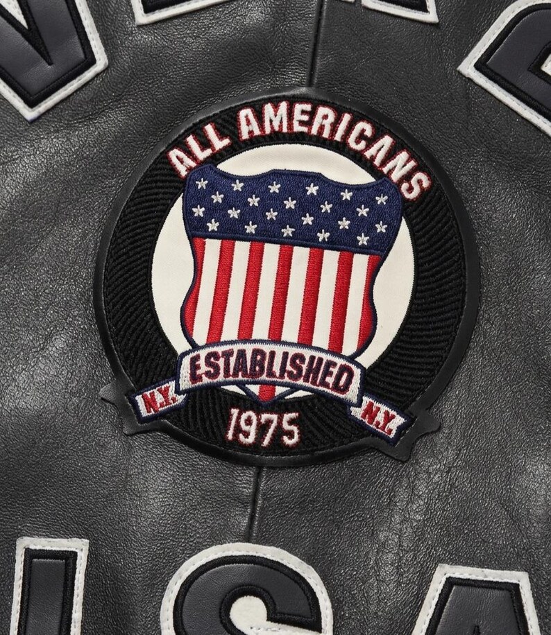 Puede incluir: Primer plano de una chaqueta de cuero negra con un parche circular. El parche presenta un dise&ntilde;o de escudo con rayas rojas y blancas, estrellas azules y las palabras "ALL AMERICANS ESTABLISHED 1975 N.Y." en letras blancas.