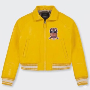 May include: A bright yellow bomber jacket with a glossy finish. The jacket features a zip-up front, a collar, and a patch on the chest with a shield design. The cuffs and hem have white stripes. The brand name "AVIREX" is visible.