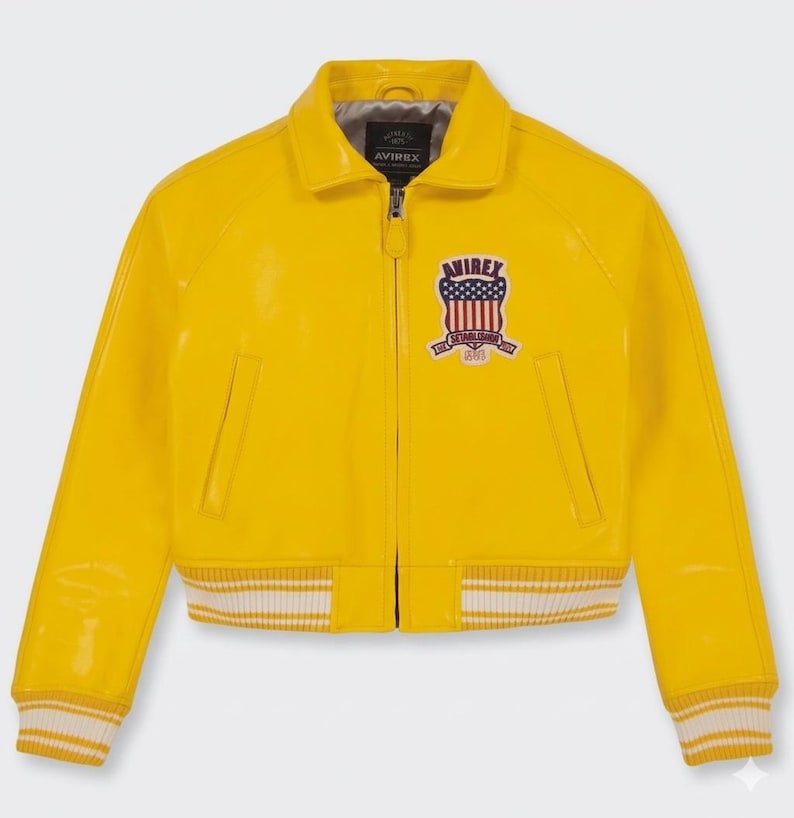 May include: A bright yellow bomber jacket with a glossy finish. The jacket features a zip-up front, a collar, and a patch on the chest with a shield design. The cuffs and hem have white stripes. The brand name "AVIREX" is visible.