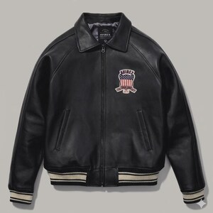 Puede incluir: Chaqueta de cuero negra con cuello, cierre de cremallera y un parche en el pecho. La chaqueta presenta un dobladillo y pu&ntilde;os acanalados con rayas blancas. El parche muestra el texto "AVIREX" y un dise&ntilde;o de bandera americana.