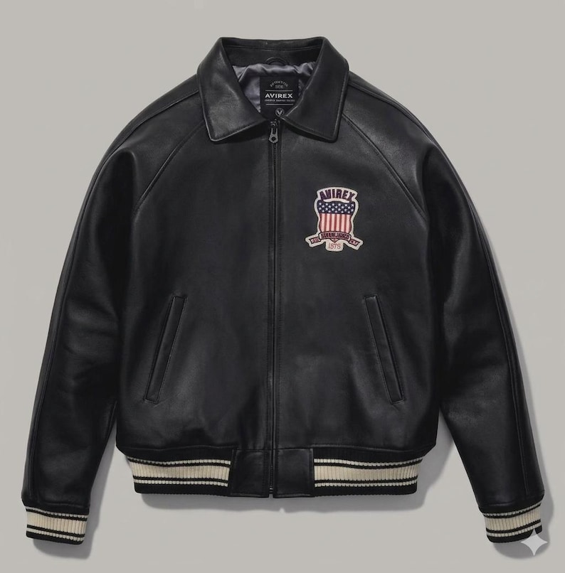 Puede incluir: Chaqueta de cuero negra con cuello, cierre de cremallera y un parche en el pecho. La chaqueta presenta un dobladillo y pu&ntilde;os acanalados con rayas blancas. El parche muestra el texto "AVIREX" y un dise&ntilde;o de bandera americana.