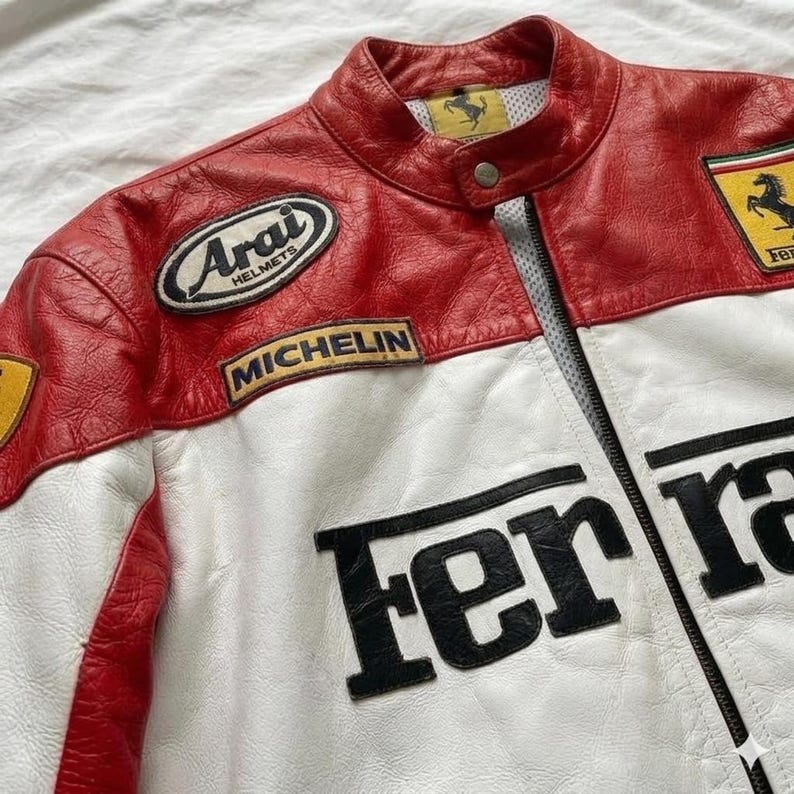 May include: Red, white, and black leather jacket with the Ferrari logo. The jacket has a stand-up collar, a zipper closure, and patches from Arai Helmets and Michelin. The Ferrari logo is on the chest and the back of the jacket.
