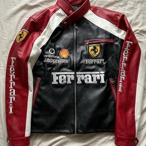 Puede incluir: Chaqueta de cuero negra, roja y blanca con el logotipo de Ferrari y otras marcas. La chaqueta tiene cremallera, bolsillos laterales y la palabra "Ferrari" bordada en la parte delantera y las mangas. Cuello alto.