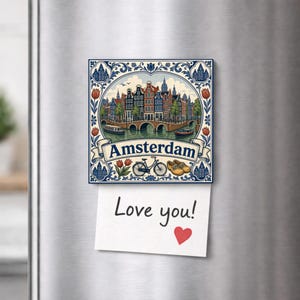 Op de afbeelding: Een vierkante decoratieve magneet met een illustratie van Amsterdam met grachtenpanden, een brug, een fiets en houten klompen. De tekst Amsterdam staat op een banner. Een wit briefje met de handgeschreven tekst Love you! en een rood hartje is bevestigd.
