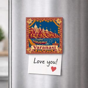 Peut inclure: Un aimant de réfrigérateur carré présentant une illustration colorée des ghats de Varanasi, un bateau et une lampe diya allumée. Le mot Varanasi est imprimé sur une bannière en bas. Une note blanche avec le texte manuscrit Love you! et un cœur rouge est fixée.