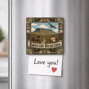 Op de afbeelding: Een vierkante koelkastmagneet met een landschapsillustratie van de Brecon Beacons met schapen, een paard en een roofvogel. De rand heeft een ruitpatroon en de tekst Brecon Beacons. Een wit briefje met Love you! en een rood hartje is bevestigd.