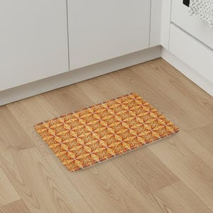 Può includere: Un tappetino rettangolare caratterizzato da un motivo geometrico ripetitivo di forme floreali a quattro petali nei colori arancione, giallo e marrone, separate da sottili linee bianche a griglia, creando un design visivo strutturato e simmetrico.