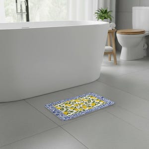 Op de afbeelding: Een rechthoekige badmat met een centraal patroon van gele citroenen en groene bladeren op een witte achtergrond, omringd door een decoratieve blauw-witte bloemenrand. De mat ligt op een grijze tegelvloer in een badkamer.