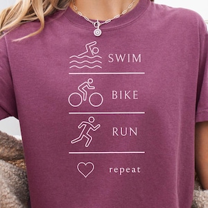 May include: A maroon t-shirt with white text and graphics. The text reads "SWIM, BIKE, RUN, repeat" with corresponding line drawings of a swimmer, cyclist, runner, and a heart. The shirt is a casual style.