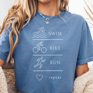 May include: A blue short-sleeved t-shirt with white text and graphics. The text reads "SWIM, BIKE, RUN, repeat" with corresponding line drawings of a swimmer, cyclist, runner, and a heart. The shirt is worn by a person.