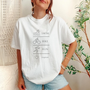May include: White t-shirt with a black graphic design. The design features the words "swim, bike, run, repeat" with corresponding illustrations of a swimmer, cyclist, runner, and a heart. The shirt is worn with blue denim shorts.